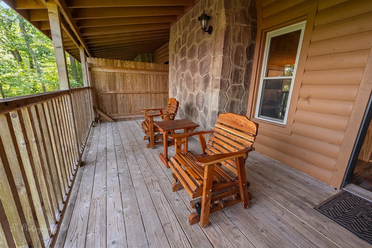 An exterior porch scene featuring wooden rocking chairs, small tables, rustic stone wall, and wooden railings. Surrounded by nature, the porch looks out onto a tranquil landscape—perfect for unwinding and recounting the day's memories.