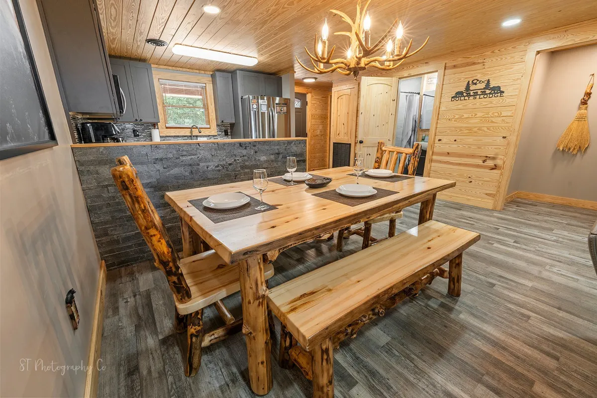 A warm, inviting dining space with a sturdy wooden dining table, chandelier, and kitchen cabinetry visible in the background. The scene reflects a welcoming atmosphere for group meals and entertaining.