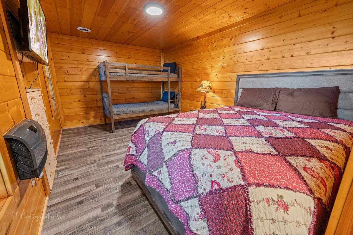 A homely bedroom showcasing a large bed with a colorful quilt and a bunk bed in the corner. The bedside lamp creates a warm, inviting ambiance, and the room feels both practical and comfortable for families or friends traveling together.