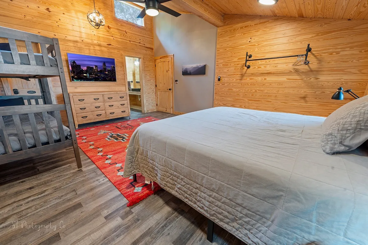 A spacious bedroom with a main bed and a twin/twin bunk, accented with a decorative rug, wall-mounted TV, and a wooden dresser. The space feels warm and comfortable, offering flexibility for couples traveling with children or close friends.