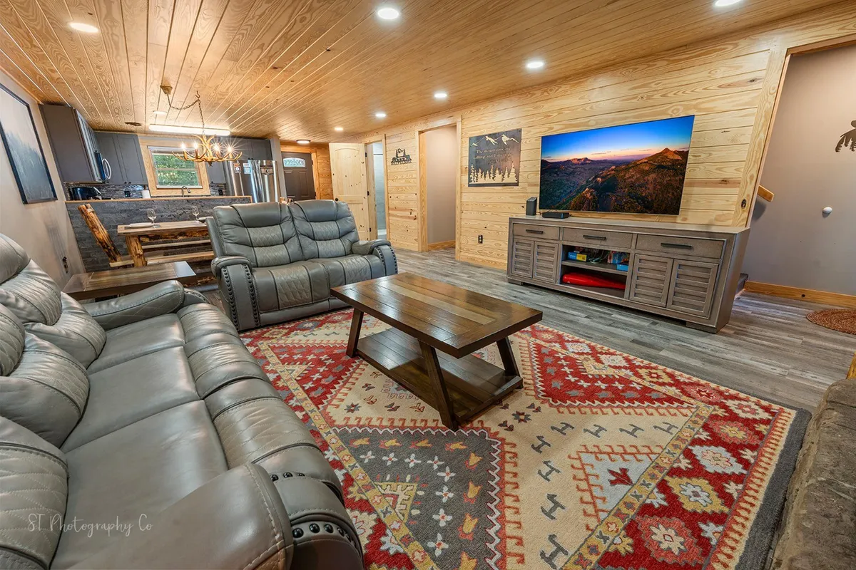 A welcoming living room with plush leather sofas arranged around a wooden coffee table. There's a decorative rug underfoot, a large television on the wall, and touches of rustic decor. The space feels warm and inviting, perfect for socializing, watching movies, or relaxing after a day in the mountains.