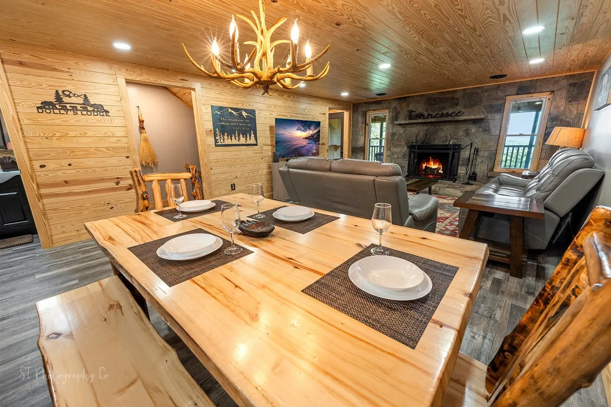 A spacious dining area featuring a wooden table set for eight, a statement chandelier overhead, and a stone fireplace in the background. The room is accented with warm wooden tones and decorative details, offering an inviting spot for family meals, game nights, or intimate gatherings.