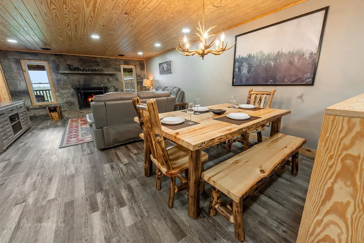 This image provides a view of a rustic wooden dining table adjacent to a comfortable gray sofa, with a prominent stone fireplace and artwork. The setting is warm and inviting, suitable for relaxation or using digital guest support services.