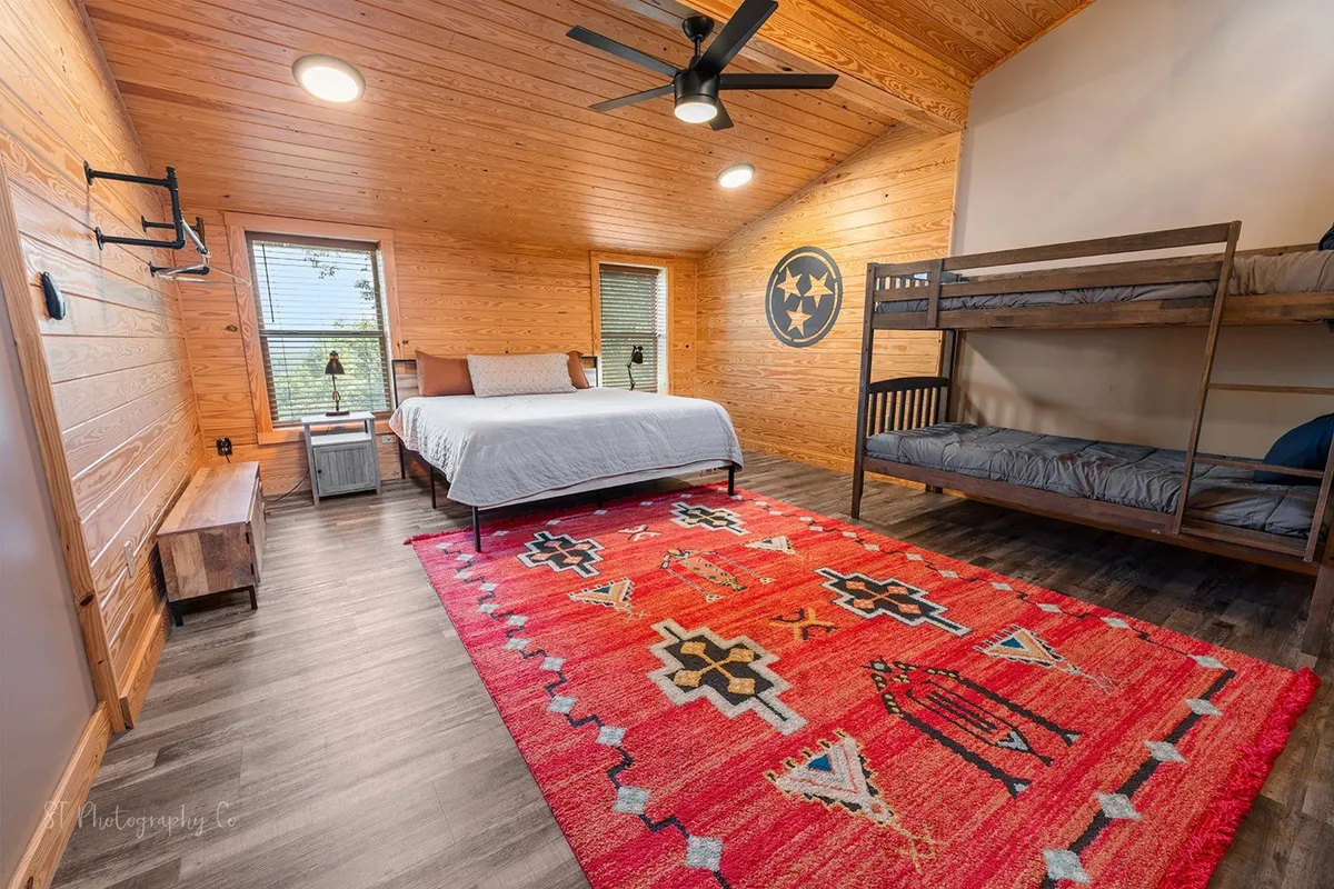 This image presents a welcoming bedroom with a main bed and twin/twin bunk beds, area rug, wood paneling, and decorative wall art. The warm woodsy accents and spacious layout make it ideal for families or groups, reflecting the comfort and communal spirit described in the section.